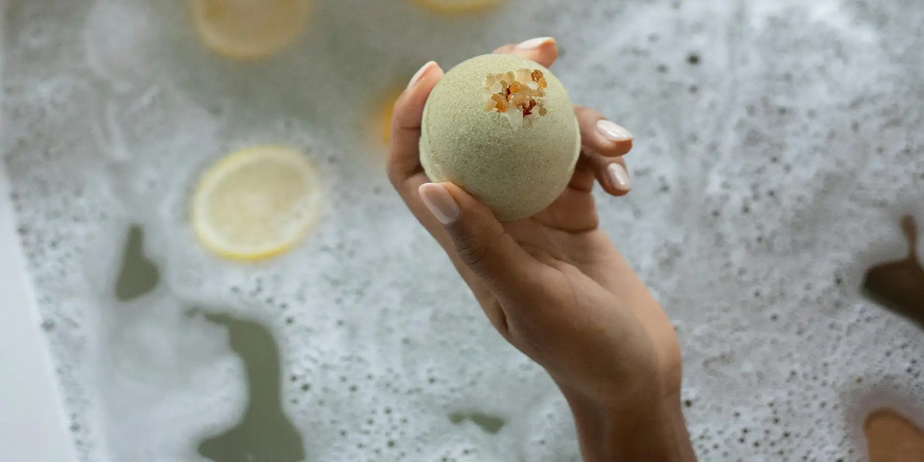 Frauenhand hält eine Badekugel über einem eingelassenen Bad